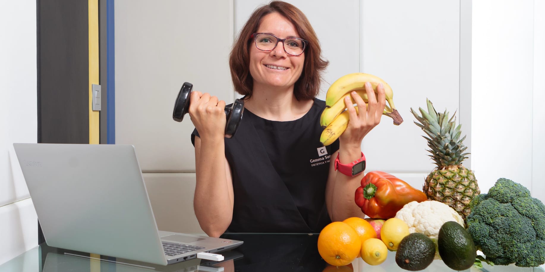 Gemma Serrano en su consulta, con una mancuerna en una mano y un plátano en la otra, junto a fruta y verdura